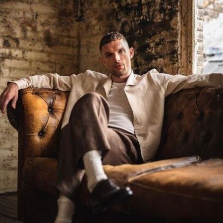 Ed Skrein is sitting on a sofa and posing with his crossed legs.